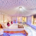 Luxurious bedroom with a carved wooden bed white linens purple pillows and a patterned ceiling and walls large window shows pink blossoms outside