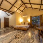 A spacious rustic bedroom with exposed wooden beams a low wooden bed marble floors and warm lighting from wall lamps a window with curtains and a ceiling fan overhead