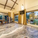 Spacious living area with a marble floor and exposed wooden ceiling beams a round wooden table with chairs a stainless fridge and a wall TV garden visible through large windows with gold curtains