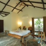 Spacious hotel room with a rustic wooden beam ceiling a large bed with white linens and green cushions and a glass dining table near a window with a leafy plant outside