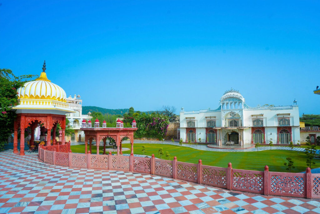 White and gold domed pavilion in a red carved courtyard with checkerboard tiles and a manicured lawn under a bright blue sky