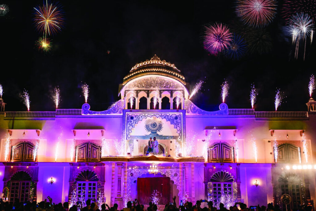 Nighttime festival at an ornate illuminated palace with colorful fireworks overhead and a crowd gathered below