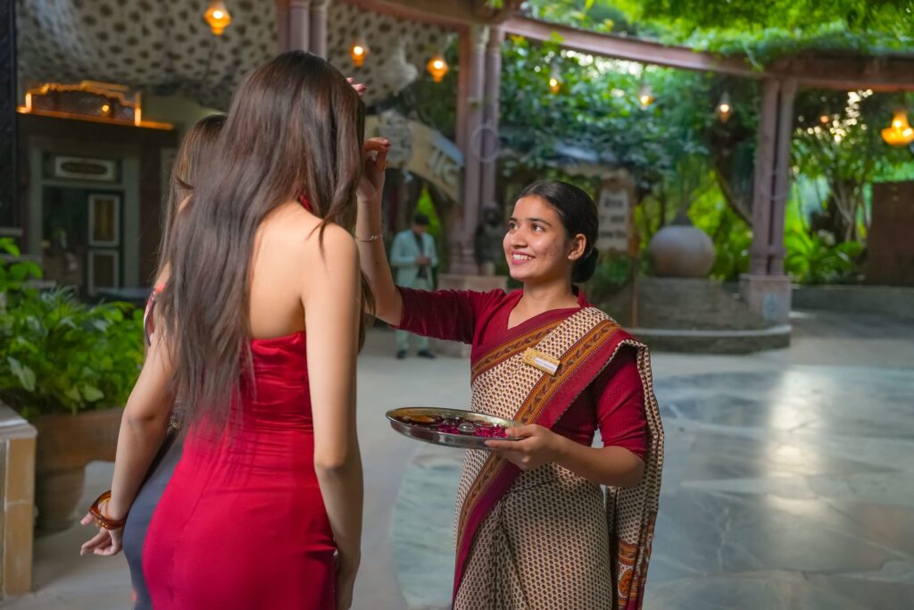 Woman in a red dress hands a silver tray of petals to another woman wearing a patterned sari in a green outdoor courtyard