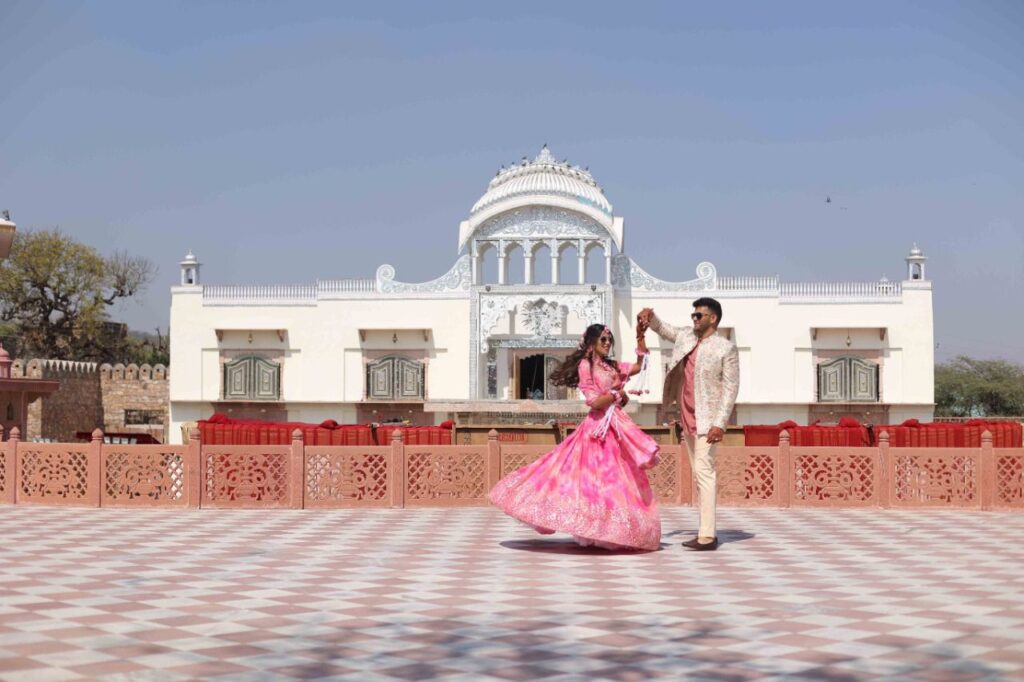 A couple in pink and cream traditional Indian outfits dancing on a checkered terrace, with an ornate white temple-like building in the background.