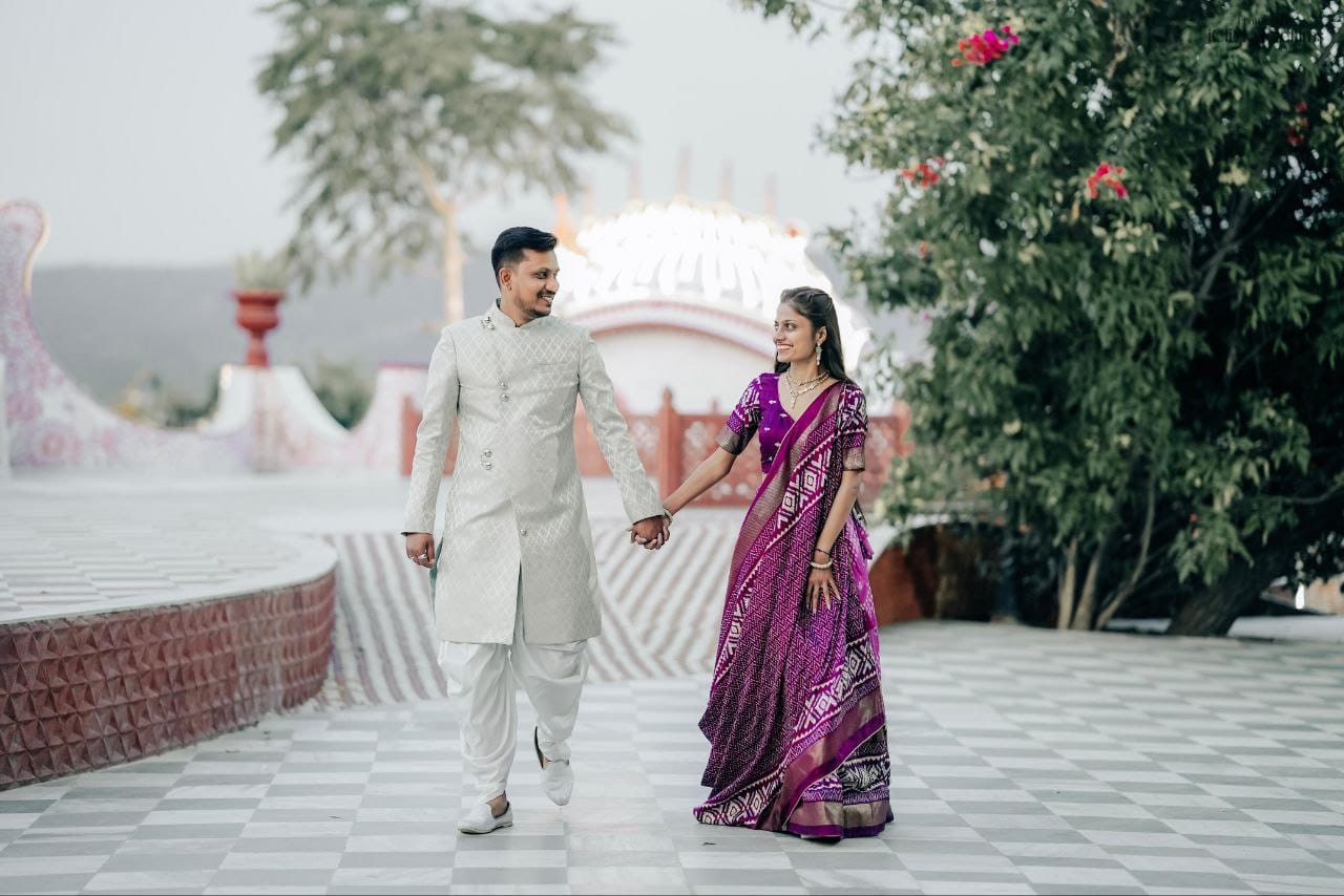 Man in a cream sherwani and a woman in a purple saree hold hands and walk together in a decorative outdoor plaza.