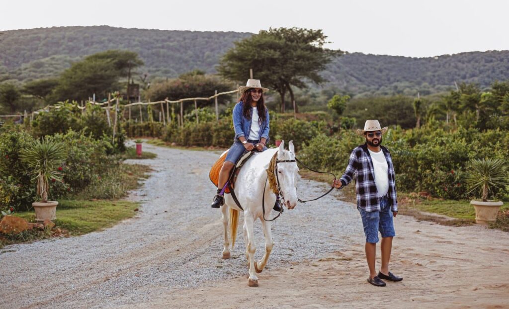 Woman wearing a hat rides a white horse while a man in a plaid shirt leads it along a gravel rural farm road.