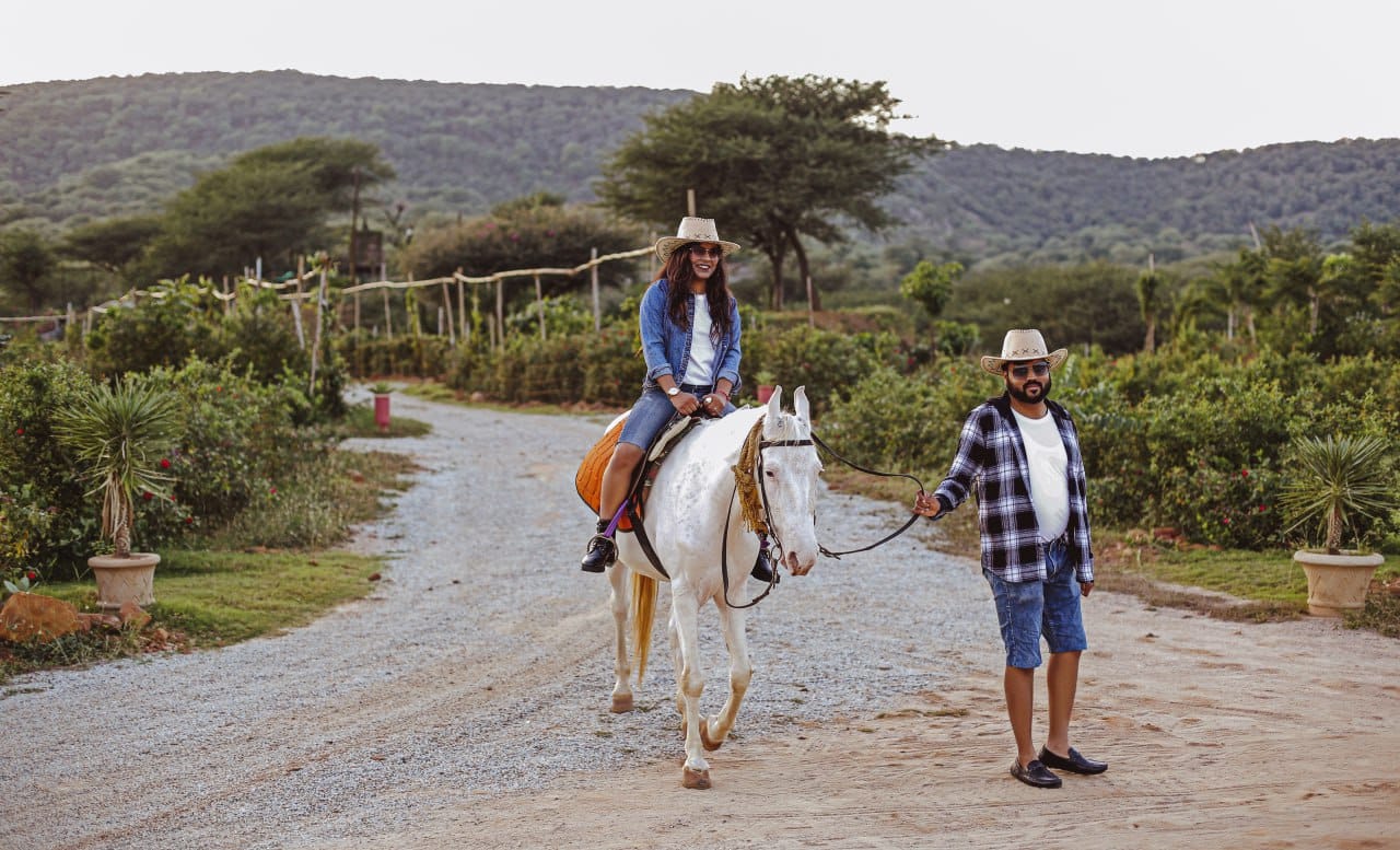 Woman rides a white horse along a gravel garden path, guided by a man in a plaid shirt holding the reins. The scene has plants and hills in the background.