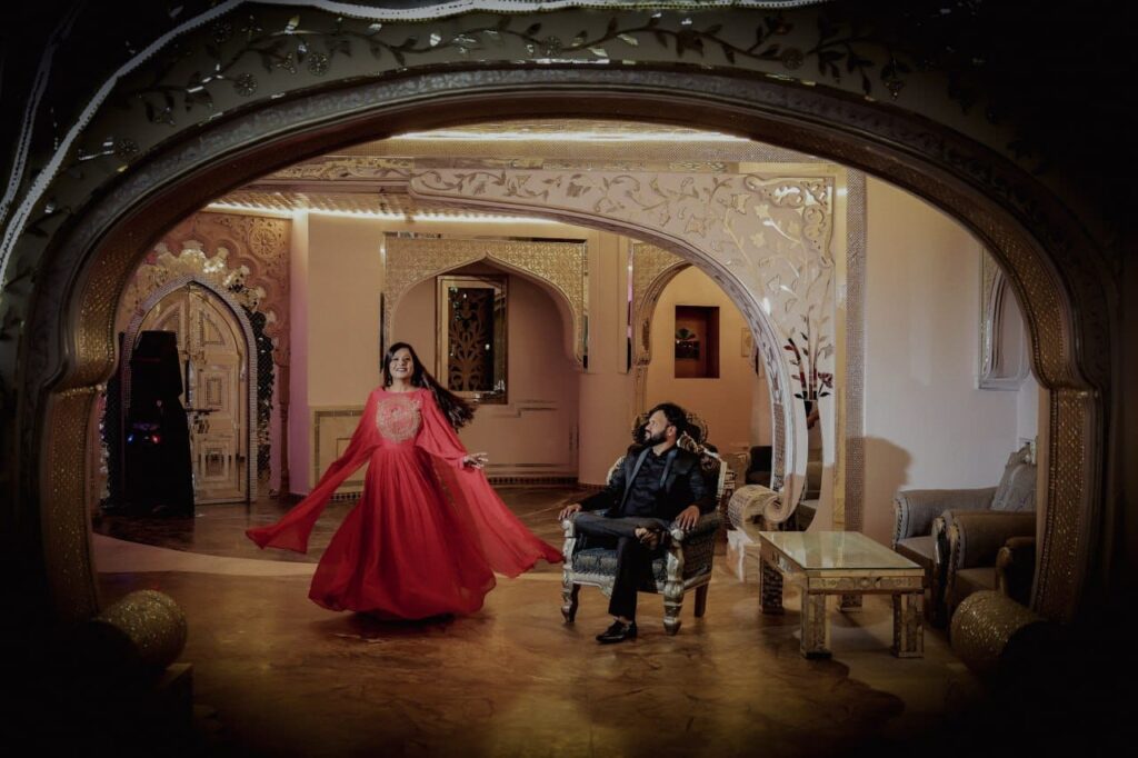 A woman in a flowing red gown dances as a man in a dark suit sits on a carved chair in an ornate, well-lit lounge.