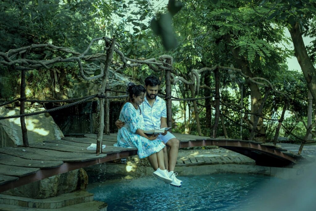 Couple sitting side by side on a rustic rope-rail bridge above a pool, reading a book together in a lush garden.