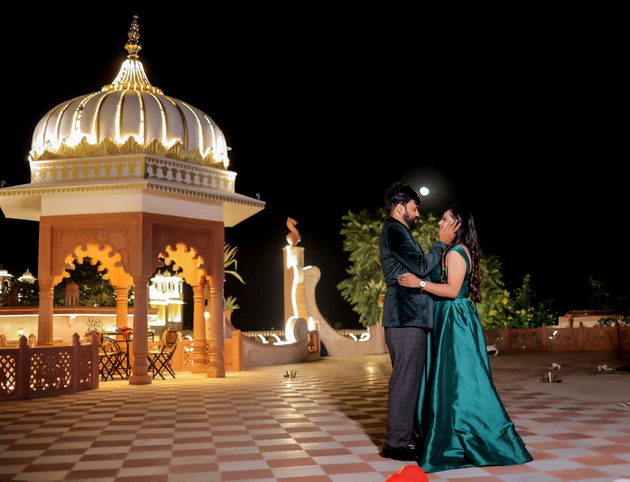 A couple in formal wear embraces on a checkered terrace at night, with a lit domed pavilion behind them.